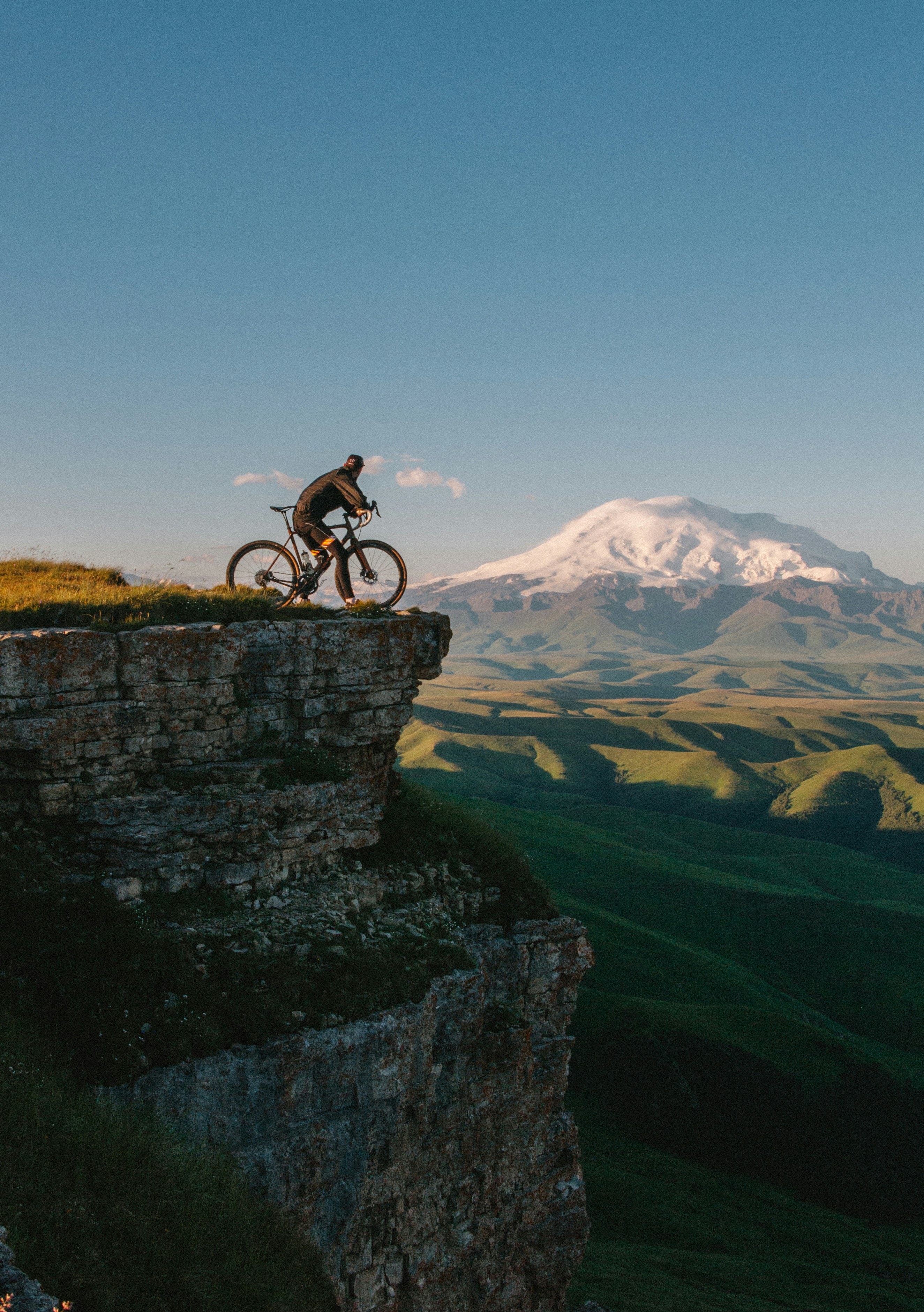 A rider stops at the edge of a high cliff, a snow-capped mountain and green valleys behind.