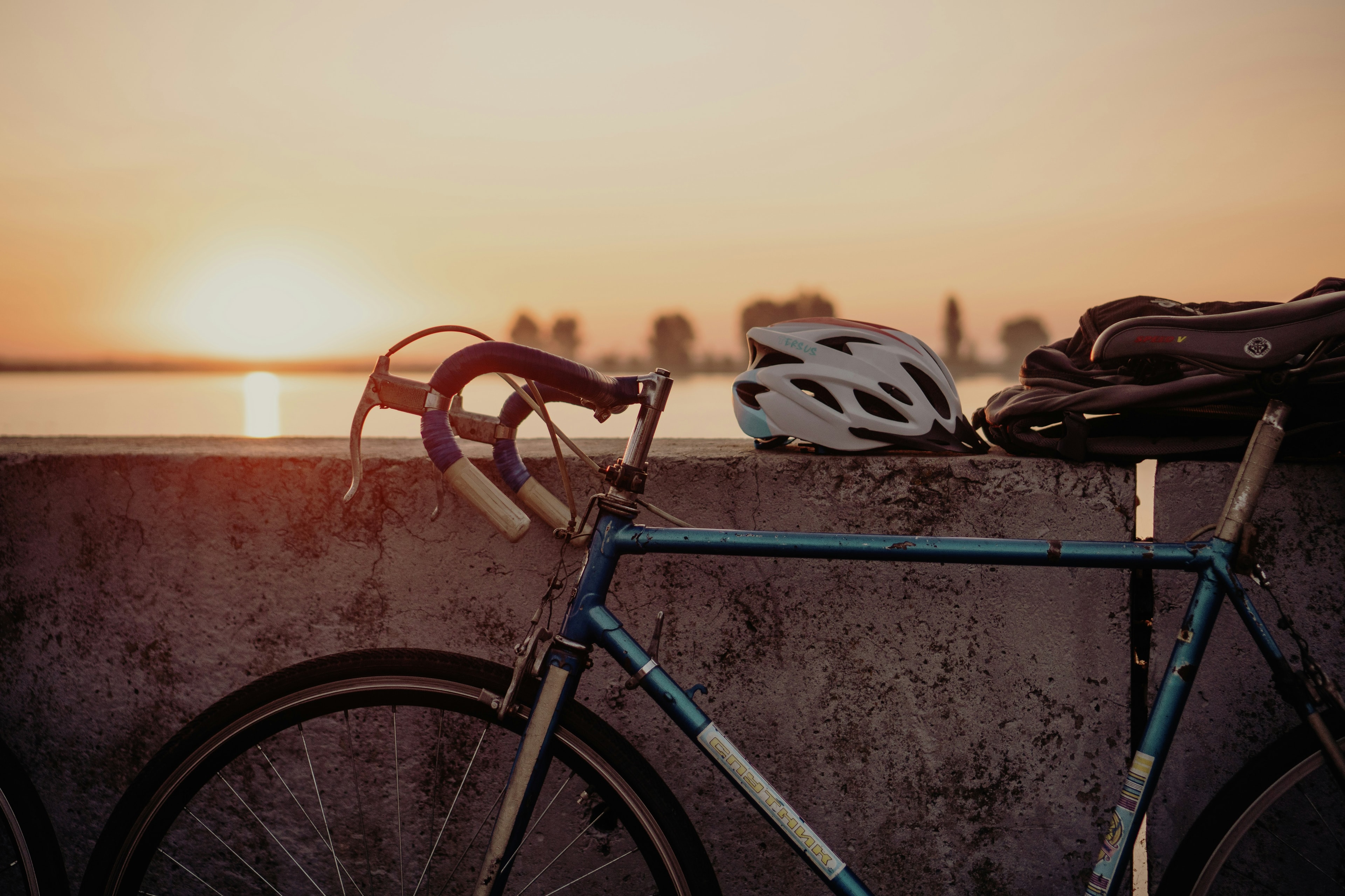 A vintage steel road bike and a helmet against a wall by the water at sunset.