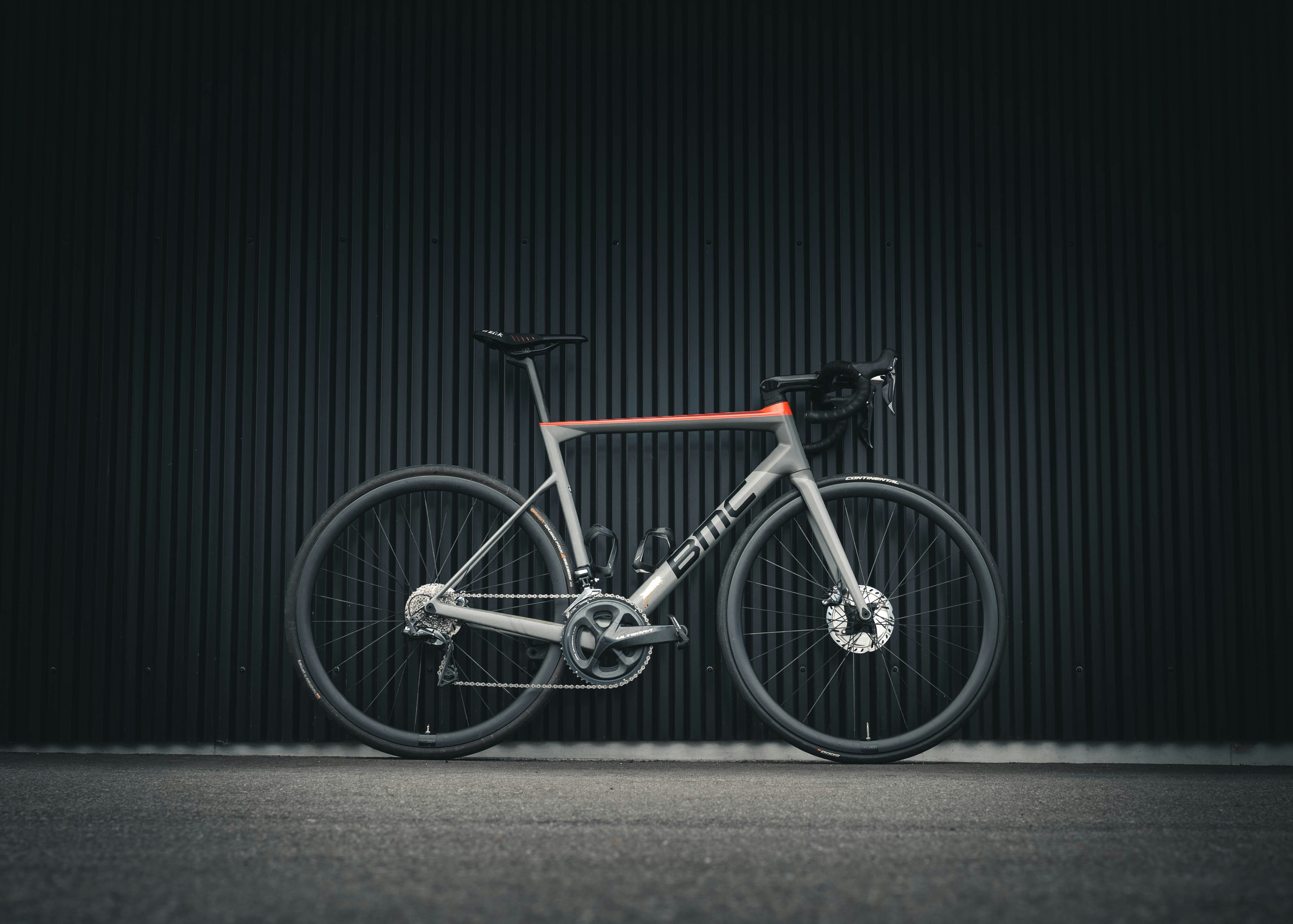 A grey and orange carbon road bike with drop bars, side-on against a dark ribbed wall.