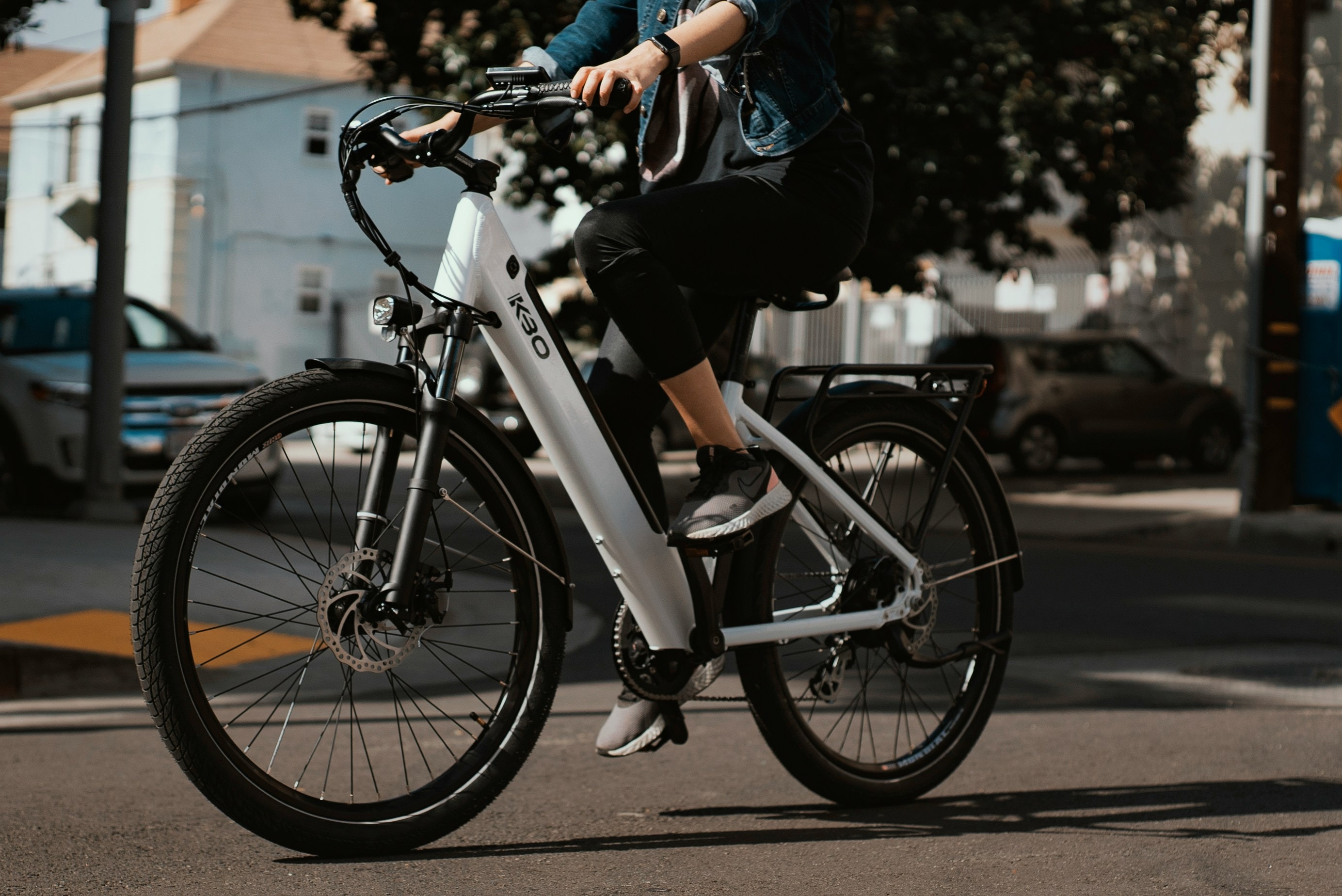 A rider pedals a white step-through E-Bike across a sunny street.