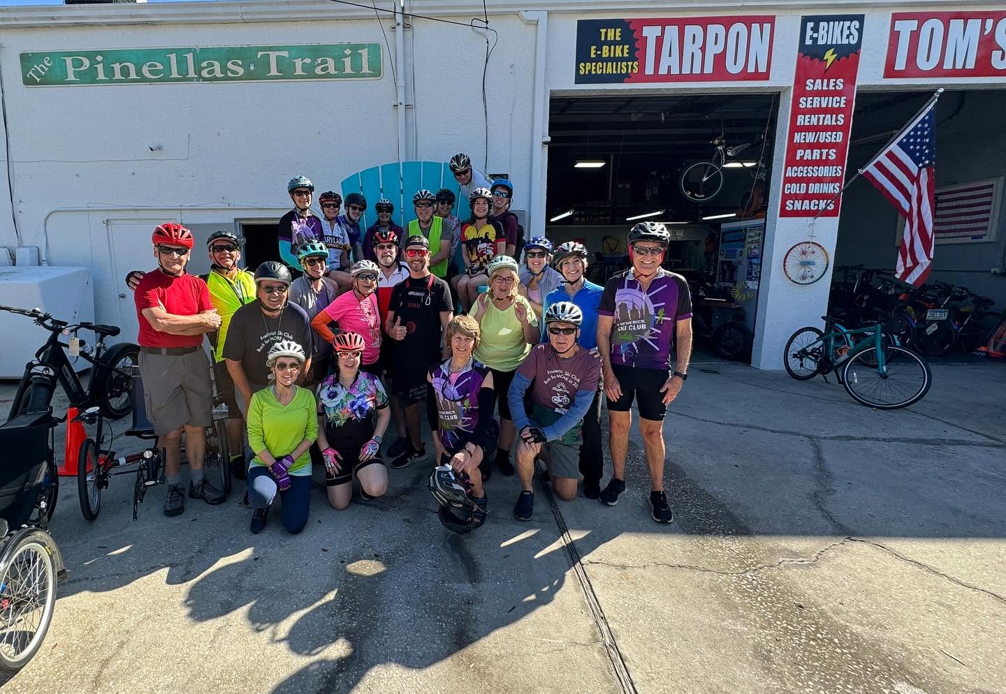 A group of riders in helmets gathers with their bikes outside Tarpon Tom’s, beneath the Pinellas Trail sign.