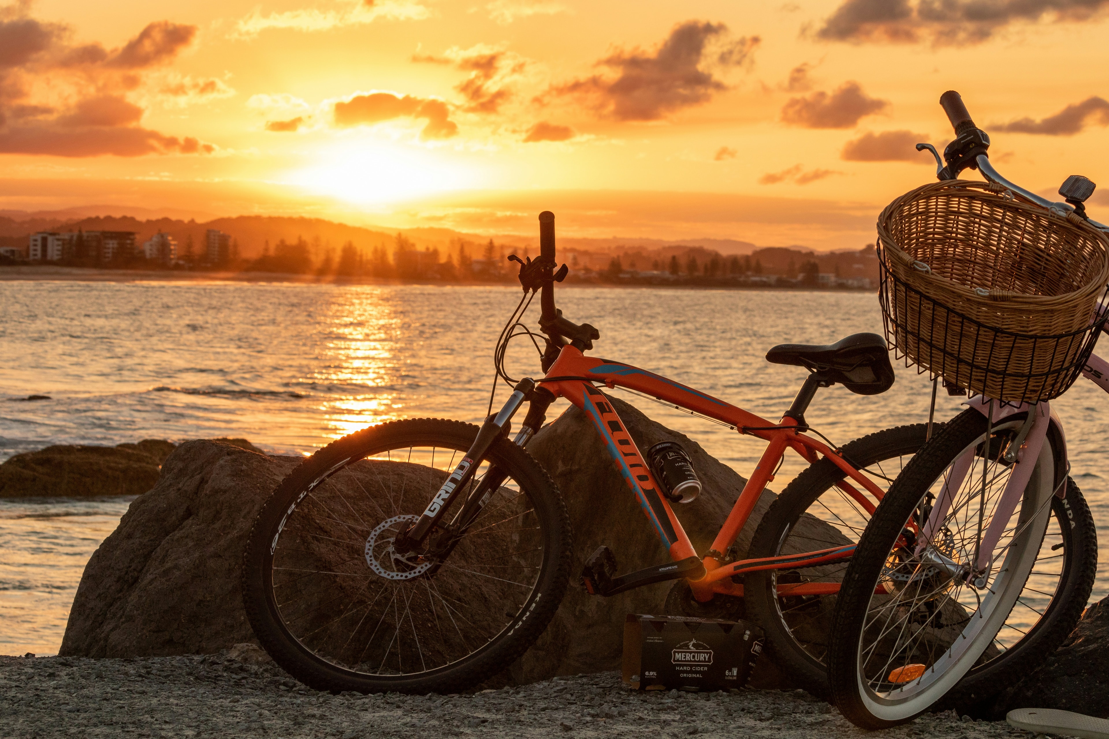 Two bikes, one with a wicker basket, rest on rocks by the water at sunset.