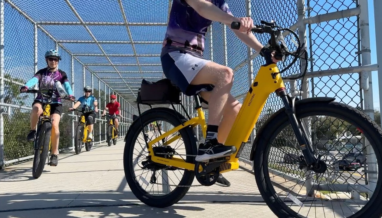 A Tarpon Tom’s tour group on yellow Velotric e-bikes crossing a fenced trail bridge on a bright day.