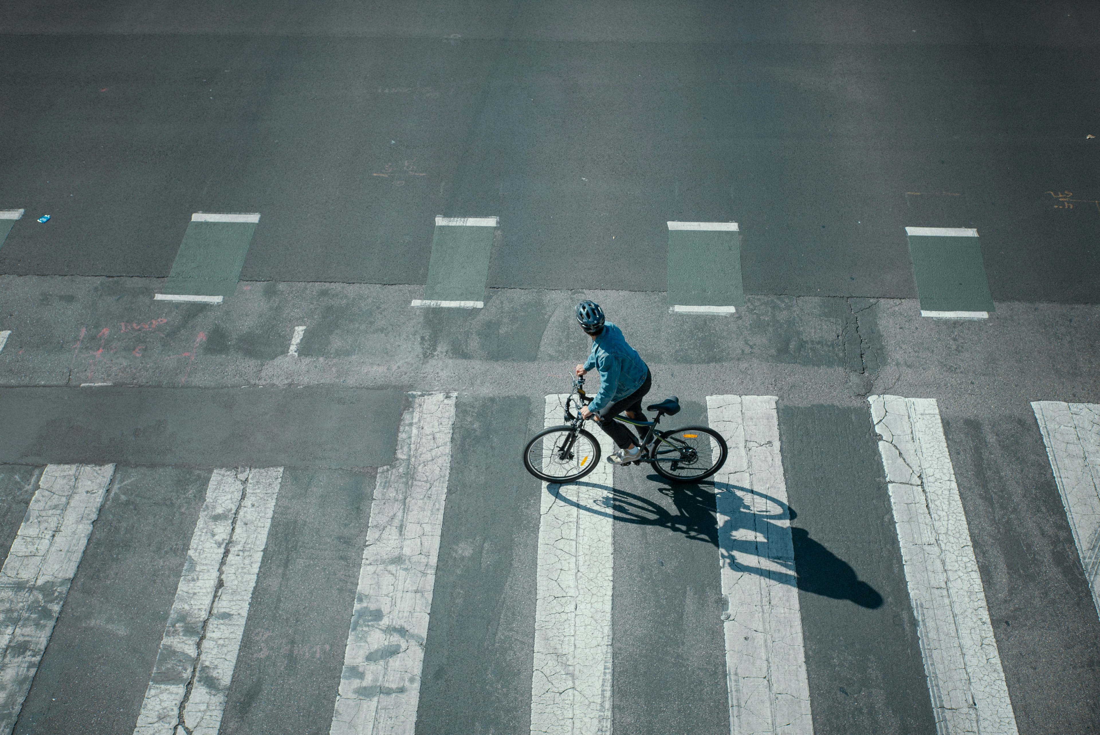 Seen from above, a helmeted rider crosses a wide striped crosswalk, a long shadow beside him.