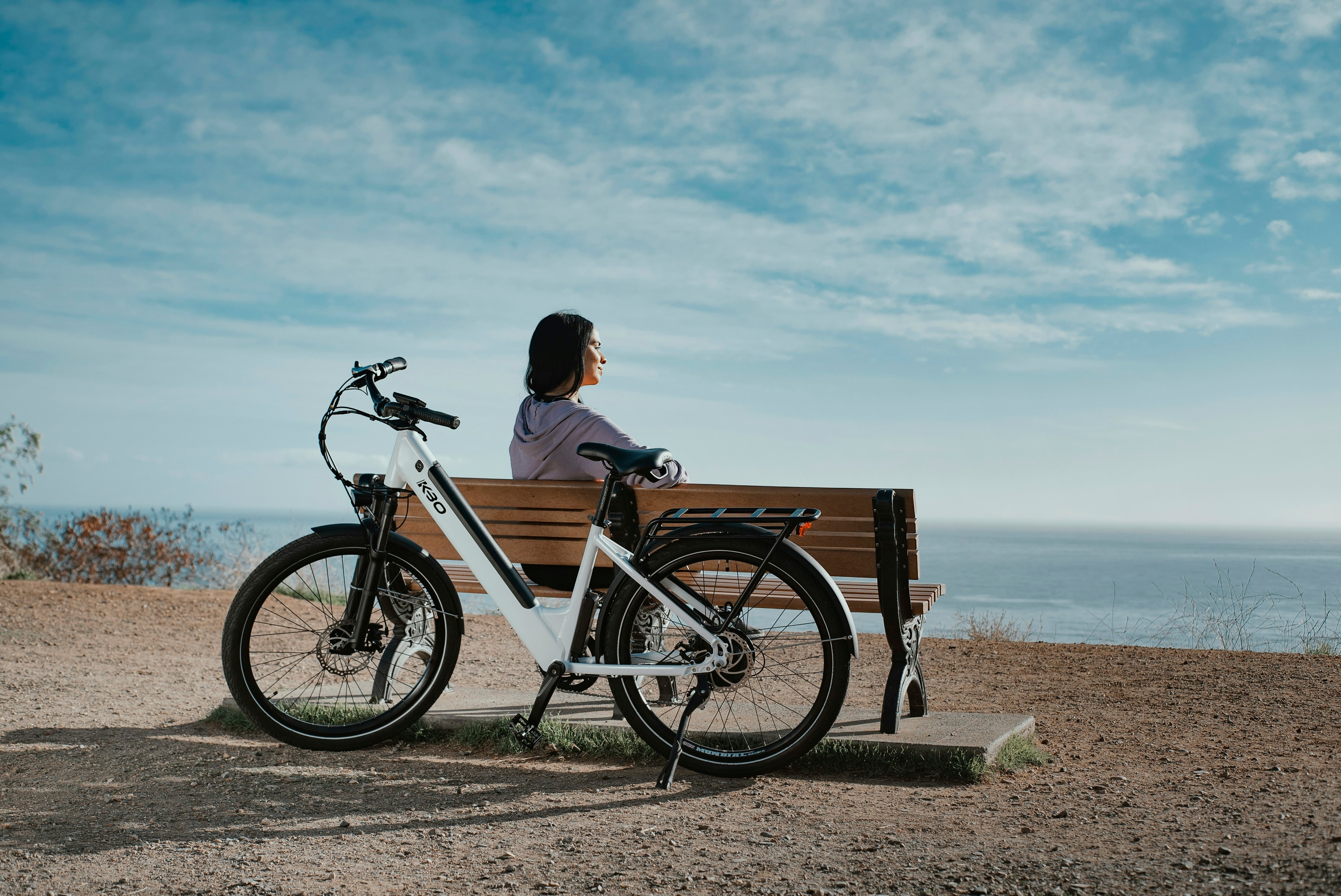 A rider rests on a bench overlooking the ocean beside a white step-through E-Bike.
