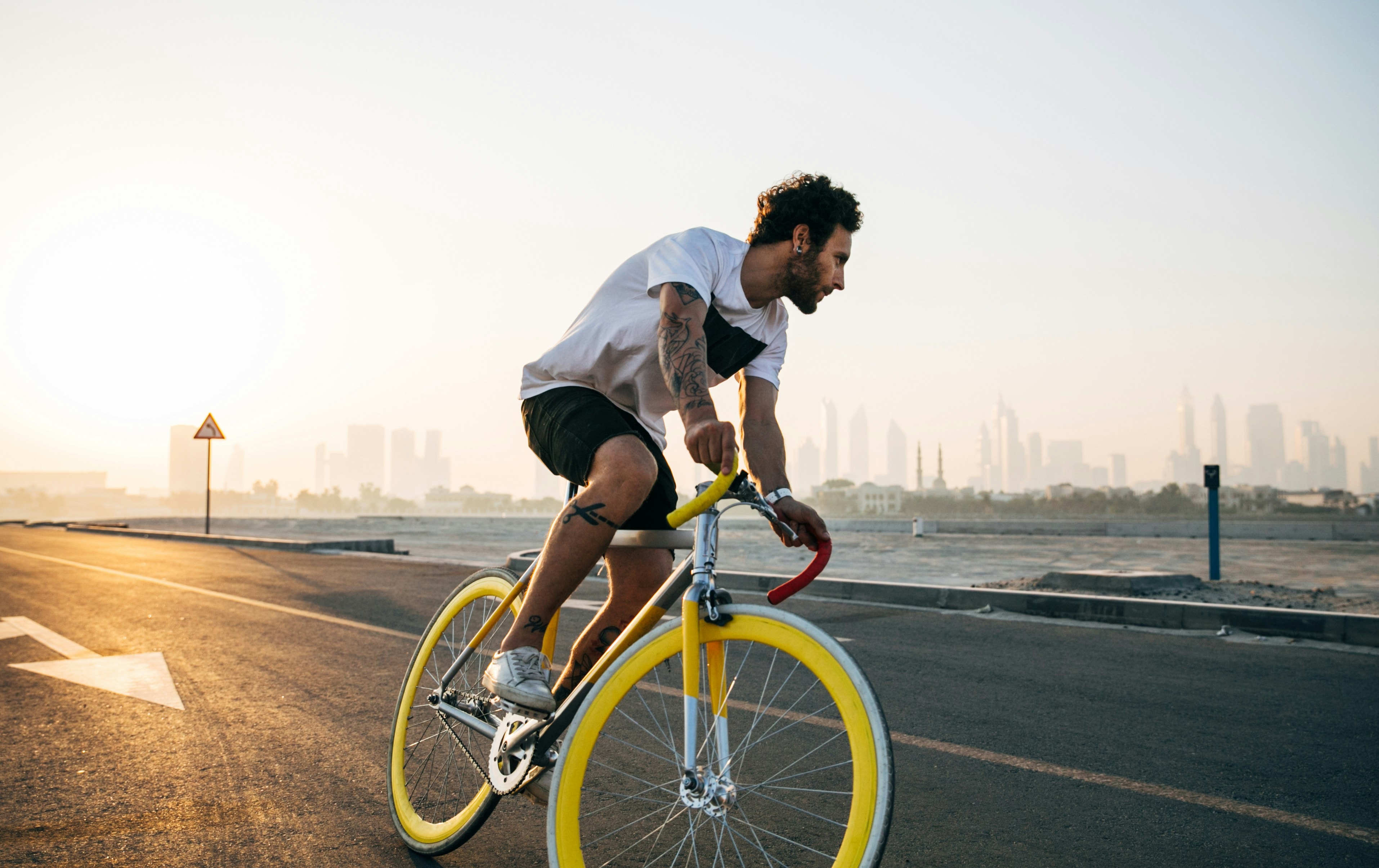A rider on a yellow fixed-gear bike leans into the road at sunrise, a city skyline in the haze behind him.