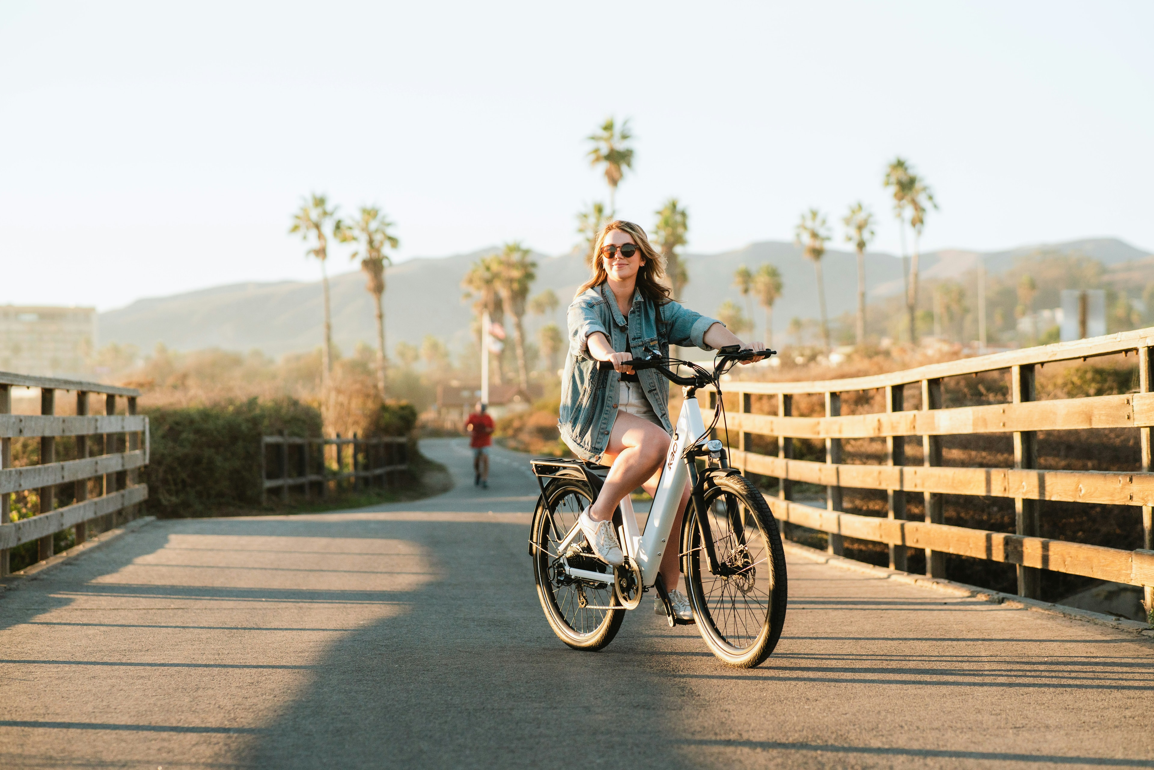 A smiling rider in sunglasses cruises a white E-Bike along a wooden boardwalk lined with palms.