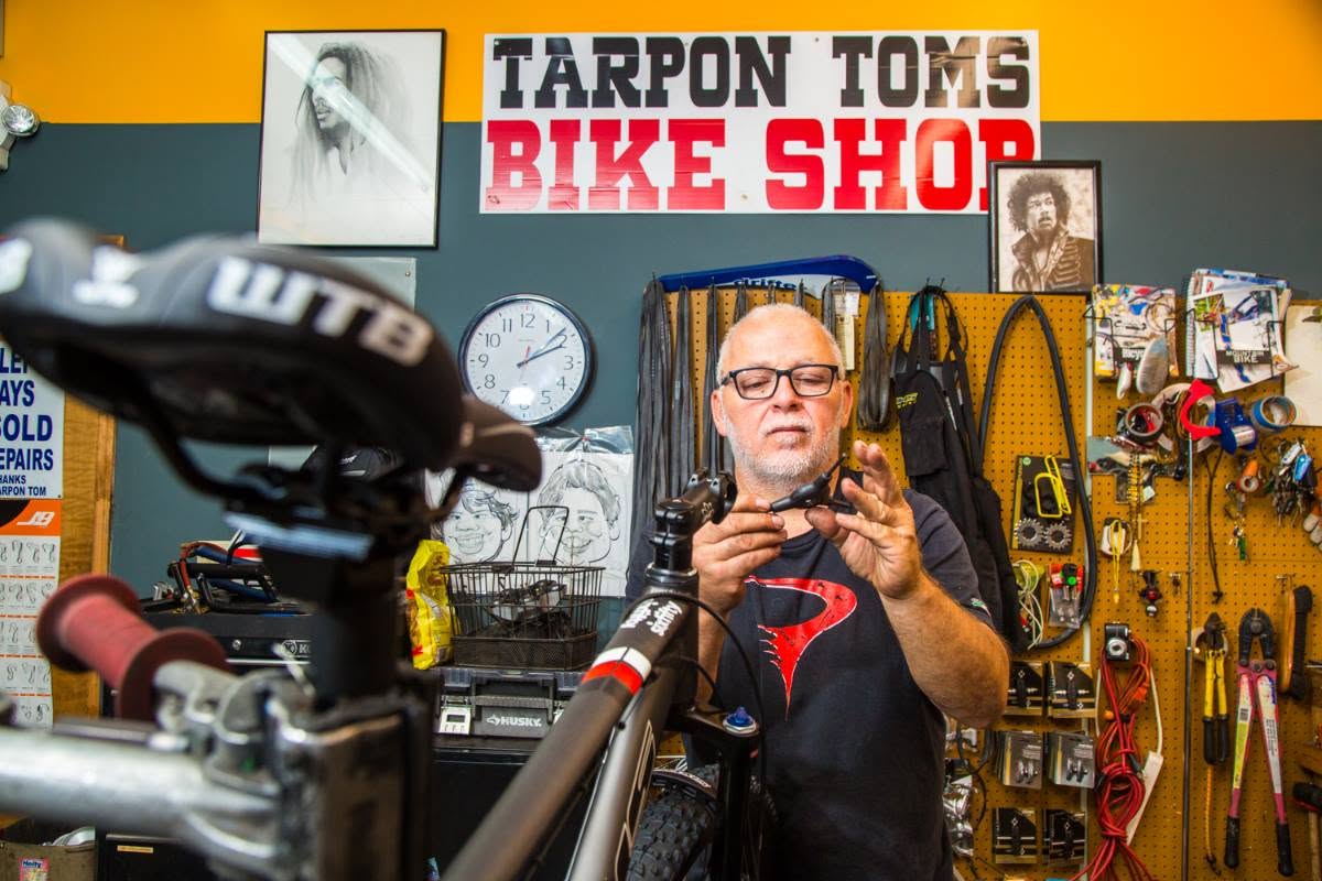 A mechanic adjusts a bike in the workshop beneath the Tarpon Toms Bike Shop sign.