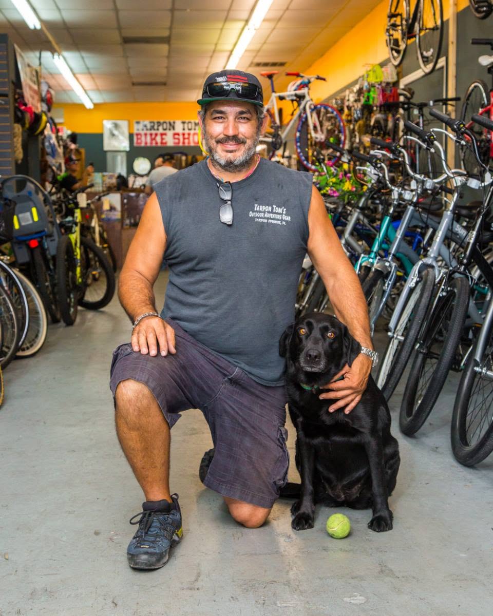 Inside Tarpon Tom’s Bike Shop in Tarpon Springs: a team member kneels beside the shop’s black Labrador among rows of bikes.